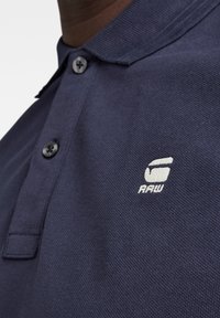 Gros plan d’un polo bleu foncé montrant deux boutons et un logo blanc brodé avec le texte "RAW" sur la poitrine.