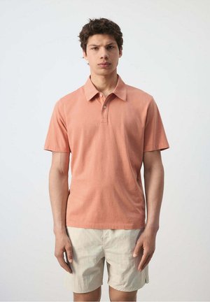 Joven con camisa polo de manga corta color rosa salmón y pantalones cortos beige claro, de pie contra un fondo liso y claro.