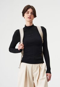 Schwarzes, geripptes Langarmoberteil mit Reißverschlusskragen, kombiniert mit lockeren, cremefarbenen Hosen und einem beigefarbenen Rucksack, der über eine Schulter getragen wird.