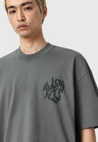 Szara, oversize bawełniana koszulka z okrągłym dekoltem i teksturowanym czarnym logo graficznym po lewej stronie klatki piersiowej. Gładkie wykończenie materiału.