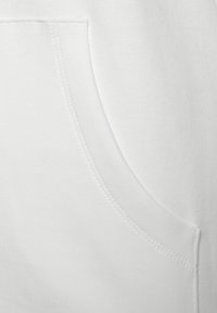 Close-up of a white fabric pocket with visible stitching on a smooth textile surface, showing pocket opening and seam details.
