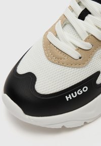 Bijele tenisice s teksturiranim mrežastim gornjim dijelom, bež zamščenim akcentima i crnim gumenim potplatom s zakrivljenim dizajnom i "HUGO" brendingom.