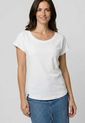 Frau mit schulterlangem braunem Haar, die ein schlichtes weißes T-Shirt mit kurzen Ärmeln und einen blauen Jeansrock trägt, leicht lächelnd.