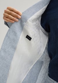 Innenansicht eines hellblauen Leinenblazers mit einem weißen gemusterten Futtermaterial und einem Etikett mit der Aufschrift "PURE LINEN". Hände halten den Blazer offen.