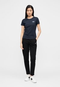 Μπλε ναυτικό t-shirt με στρογγυλή λαιμόκοψη και λογότυπο, σε συνδυασμό με μαύρο στενό παντελόνι φόρμας και λευκά αθλητικά παπούτσια με μαύρες λεπτομέρειες.