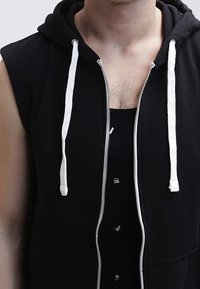 Sudadera sin mangas negra con cremallera y cordones blancos, presenta una cremallera frontal y una camiseta de tirantes negra con pequeños patrones blancos debajo.