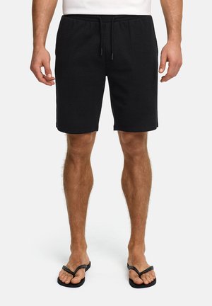 Schwarze, strukturierte Shorts in Knielänge mit Kordelzug, getragen von einem Mann, der barfuß in schwarzen Flip-Flops vor einem weißen Hintergrund steht.