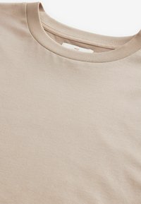 Beige színű, kerek nyakú póló puha pamut anyagból. Simább textúrával rendelkezik, egyszerű dizájnnal és bordázott gallérral. Nincsenek rajta minták vagy díszítések.