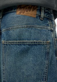 Farkut haalistuneessa sinisessä sävyssä, joissa on teksturoitu viimeistely, nahkainen merkintä vyötäröllä ja näkyvät saumat taskujen ympärillä.