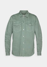 Vihreä denim-paita pitkillä hihoilla, kahdella rintataskulla, napitus edessä ja pyöristetty helma. Silkkinen pinta, jossa on hienovaraisia vaihdoksia.