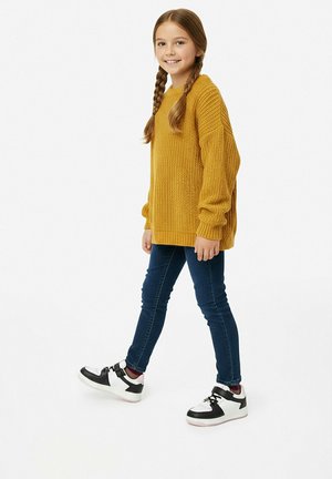 Junges Mädchen mit geflochtenem Haar, das einen senfgelben Pullover, dunkle Jeans und schwarz-weiße Turnschuhe trägt und vor einem schlichten weißen Hintergrund geht.