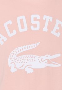 Camiseta de algodón rosa con un gran logo de cocodrilo blanco y texto en negrita que dice "LACOSTE" encima del gráfico. Textura suave.