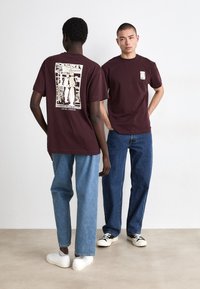 Burgunderfargede bomulls-T-skjorter med grafisk trykk på baksiden. En modell har på seg lyseblå jeans, den andre har på seg mørkeblå jeans. Hvite joggesko.