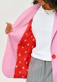 Růžový blazer s jasně červenou podšívkou a bílými srdíčkovými vzory. Pod ním bílé tričko s kulatým výstřihem a viditelné šedé pruhované kalhoty.