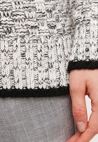 Svart och vit stickad tröja med texturerad mönster vid ärmslutet, som har en hel svartsida. Hand som håller ärmen mot grått tyg.