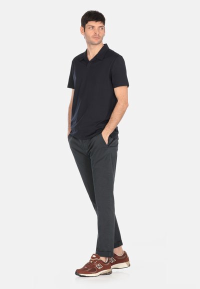 Camisa polo de manga corta azul marino, pantalones ajustados gris oscuro y zapatillas deportivas marrones con suelas blancas. Modelo de pie con las manos en los bolsillos.