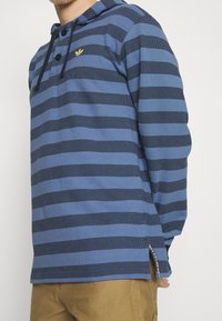 Blå och marinrandig långärmad polo med huva, knappslå och en liten gul logotyp på bröstet. Texturerat tyg.