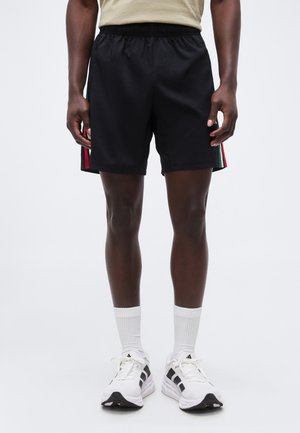 Man draagt zwarte sportshorts met rode, witte en groene strepen aan de zijkanten, witte sokken en witte Adidas sneakers, staand tegen een witte achtergrond.