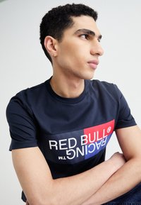 Tamsiai mėlynas marškinėliai pagaminti iš lygaus audinio, su dideliu raudonos ir mėlynos spalvos logotipu, ant kurio blokuotais rašmenimis užrašyta "RED BULL RACING."