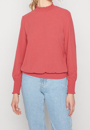 Kvinde iført en langærmet koralpink top med flæset krave og manchetter, kombineret med lyseblå jeans med høj talje, mod en hvid baggrund.