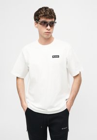 Νεαρός άνδρας με σγουρά μαλλιά φοράει αθλητικά σκούρα γυαλιά ηλίου, λευκό μπλουζάκι με μικρό λογότυπο και μαύρο παντελόνι με τσέπες, με τα χέρια στις τσέπες.