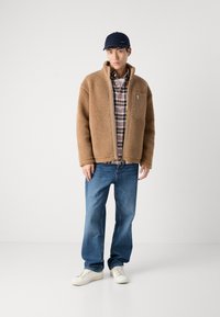 Brun fleecejakke med en lynlåslomme på brystet, parret med en ternet skjorte, blå jeans og hvide sneakers. En marineblå cap fuldender looket.