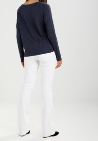 Marinblå långärmad topp med rundad halsringning, tillsammans med vita utsvängda byxor. Tyget verkar mjukt med en slät struktur. Svarta skor.