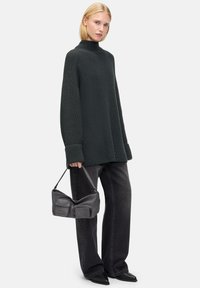 Un pull en maille vert foncé avec un col montant, port�é avec un jean noir large et un sac à main noir décontracté. Le modèle se tient devant un fond neutre.