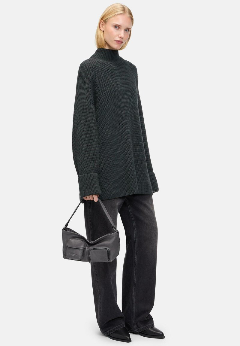 Un pull en maille vert foncé avec un col montant, porté avec un jean noir large et un sac à main noir décontracté. Le modèle se tient devant un fond neutre.