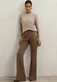 Pulover din amestec de lână bej, cu guler rotund; pantaloni largi de culoare maronie, croiți; țesătură fină; textură moale; modelul se află într-un cadru neutru.