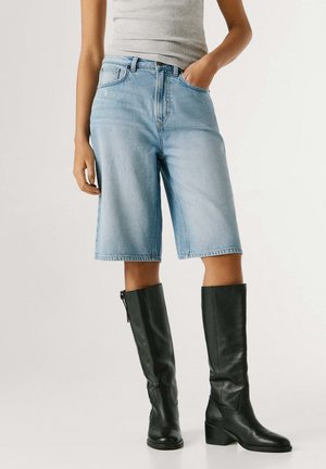 Personne portant un bermuda en denim bleu clair, un débardeur gris et des bottes en cuir noires montantes jusqu'aux genoux, debout avec une main dans la poche.