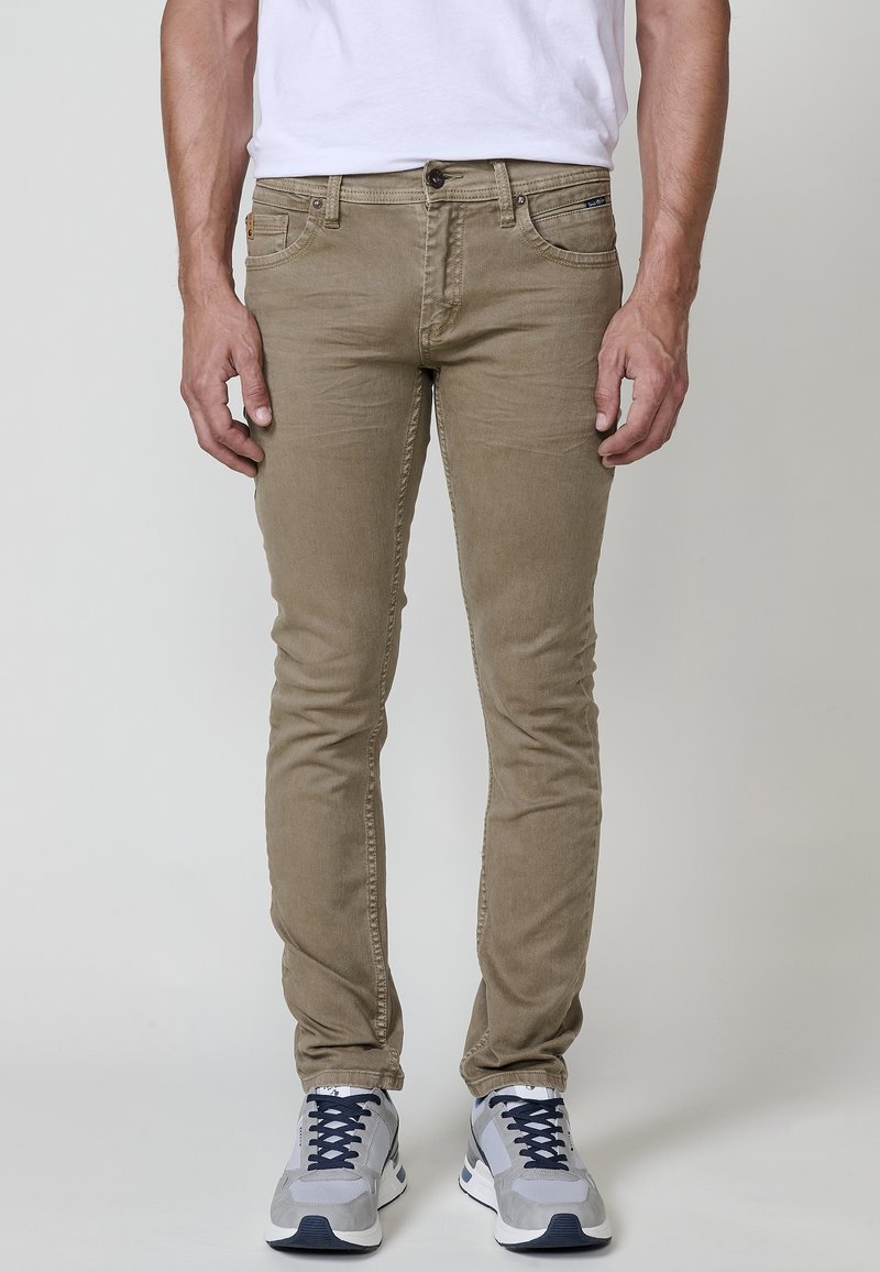 Beige skinny farmer nadrág, pamut keverékkel, öt zsebbel, övhurkokkal és gombos záródással. Szürke sportcipővel párosítva.