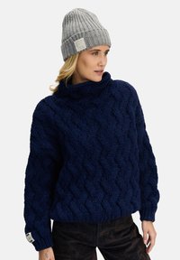 Granatowy sweter w splot kablowy z wysokim kołnierzem, teksturowanym wzorem i ściągaczami na mankietach. Dobraną do niego jasnoszarą, prążkowaną czapkę z dzianiny.