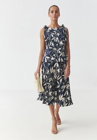 Top navy cu imprimeu floral și bretele volănașe, asociat cu o fustă asortată. Material neted, design floral alb și tiv evazat. Poșetă bej și pantofi cu toc.