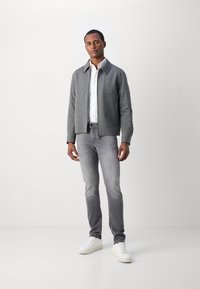 Chaqueta gris con cremallera y cuello, combinada con jeans gris claro y zapatillas blancas. La tela parece texturizada.