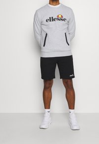 Hombre vistiendo un sudadero gris de ellesse, pantalones cortos negros de ellesse, calcetines blancos de Nike y zapatillas blancas, de pie contra un fondo claro y liso.