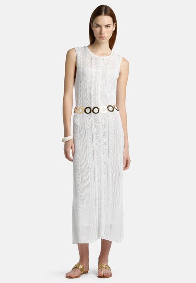 Vestido maxi de punto blanco con un patrón texturizado, que presenta un cinturón ancho de círculos entrelazados en oro y plata, combinado con sandalias planas.