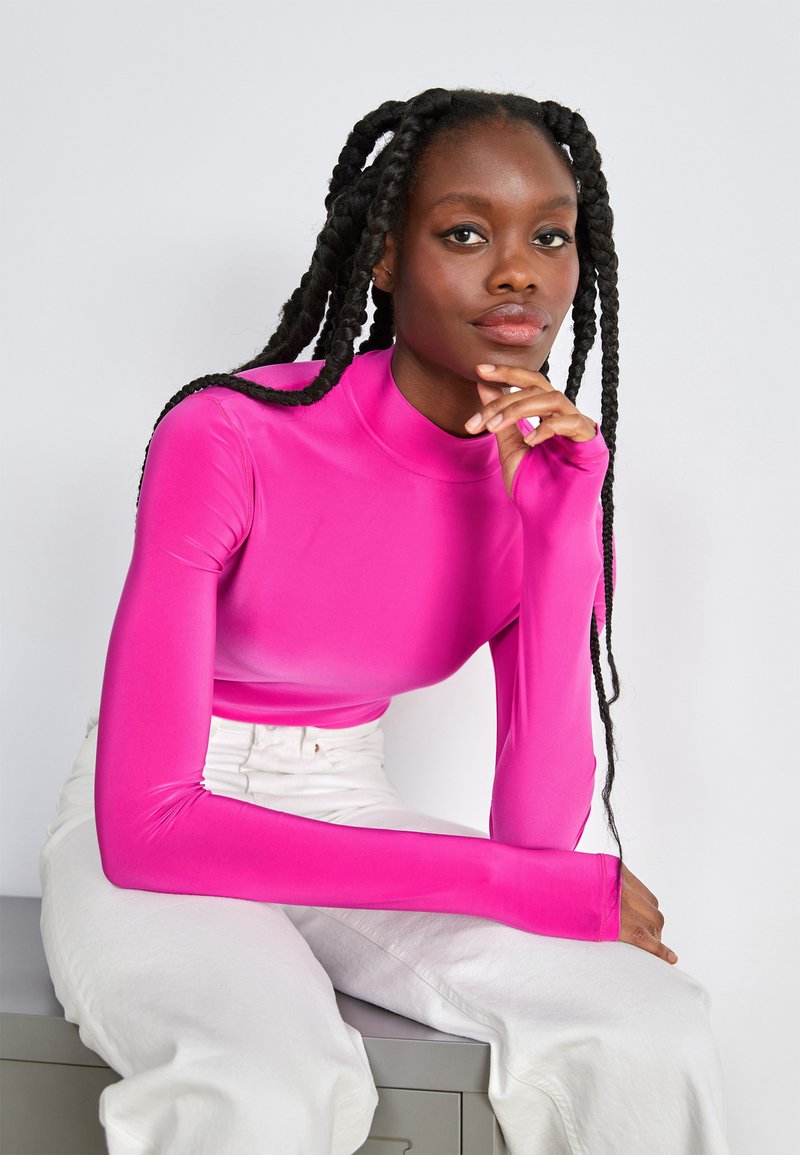 Haut ajusté à manches longues de couleur rose vif, fabriqué dans un matériau lisse et extensible avec un col haut. Porté avec un pantalon blanc.