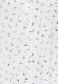 Tissu blanc avec un petit motif floral bleu et des feuilles vert clair, montrant une rangée de boutons blancs au centre.