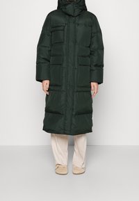 Mørkegrøn, polstret lang jakke med lynlås og knapper, der har flere frontlommer og en høj krave. Blød tekstur, afslappet pasform.