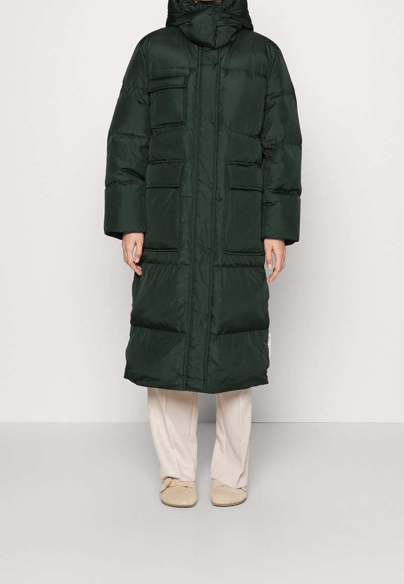 Mørkegrøn, polstret lang jakke med lynlås og knapper, der har flere frontlommer og en høj krave. Blød tekstur, afslappet pasform.