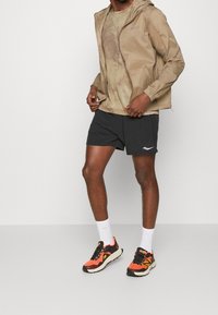 Světle hnědá zipová mikina s kapucí přes béžové vzorované tričko, doplněná černými šortkami a vícebarevnými běžeckými botami. Bílé kotníkové ponožky.