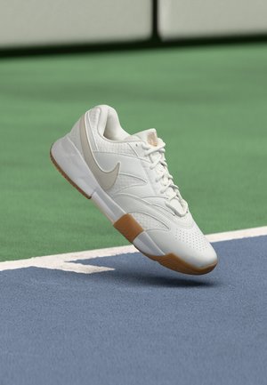 Biely Nike teniska s gumovou podrážkou vznášajúca sa nad modro-zeleným povrchom tenisového kurtu blízko bielej čiary.
