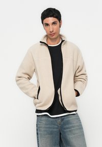 Beige fleecejakke med høy krage og svarte glidelåser, med sidelommer og kontrasterende svarte detaljer, båret over en svart skjorte.