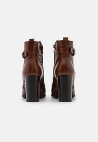 Hnědé kožené kotníkové boty s texturovaným vzorem, se dvěma bočními přezkami a masivním černým vysokým podpatkem. Hladký a lesklý povrch.