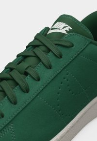 Κοντινή λήψη ενός πράσινου αθλητικού παπουτσιού Nike με κορδόνια στο ίδιο χρώμα, λευκό λογότυπο στη γλώσσα και διατρήσεις στα πλευρά σε λευκή σόλα.