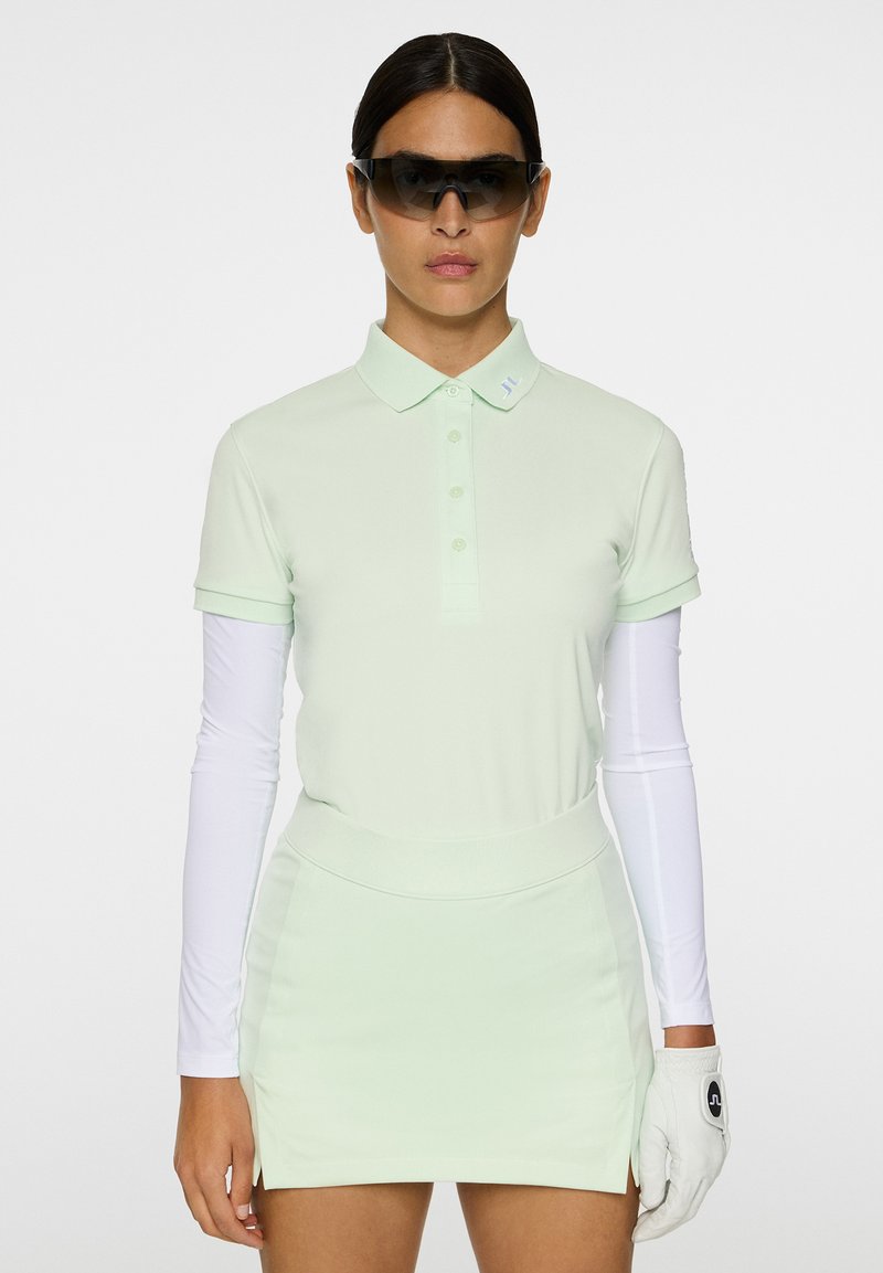 Vihreä vaaleanpuhdas polo-paita, jossa on lyhyet hihat ja napitettavat kaulukset, yhdistettynä vastaavaan tiivistettyyn hameeseen, valkoisten pitkähihasten päällä ja golf-käsine.