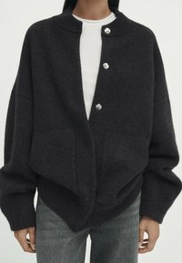 Donkergrijze oversized cardigan van zachte wol met een ronde kraag, vier zilverkleurige knopen en voorzakken. Draag over een wit shirt.