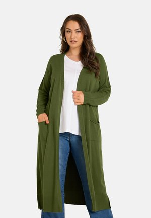 Femme portant un long cardigan vert olive par-dessus un top blanc et un jean bleu, debout avec une main dans la poche et l'autre tenant le cardigan.