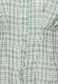 Tecido de camisa xadrez verde e branca com uma textura suave, apresentando uma fileira de botões e pregas subtis ao longo das costuras.